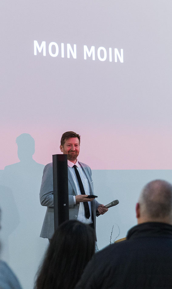 Peter Bossecker smirking while giving a talk, the words 'Moin Moin' on a slide in the background.